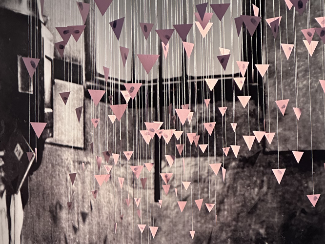 Kunstinstallation mit herabhängenden Fäden und dreieckigen Pappelementen in Rosatönen vor einem schwarz-weißen Fotohintergrund.