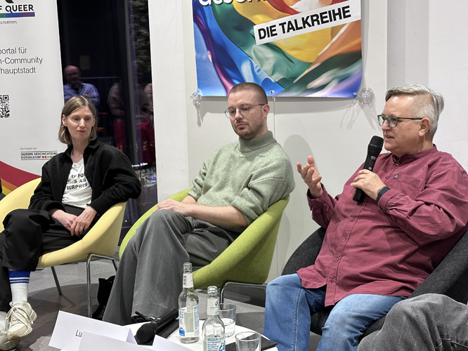 Drei Personen bei einer Podiumsdiskussion, die rechte Person hält ein Mikrofon, im Hintergrund ein regenbogenfarbenes Banner.