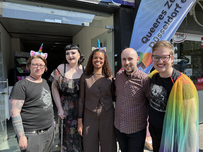 Fünf Personen stehen lächelnd vor dem Eingang des Queeres Zentrum Düsseldorf. Zwei tragen Katzenohren, eine eine Regenbogenflagge als Cape. Im Hintergrund ist ein Banner mit dem Namen des Zentrums zu sehen.