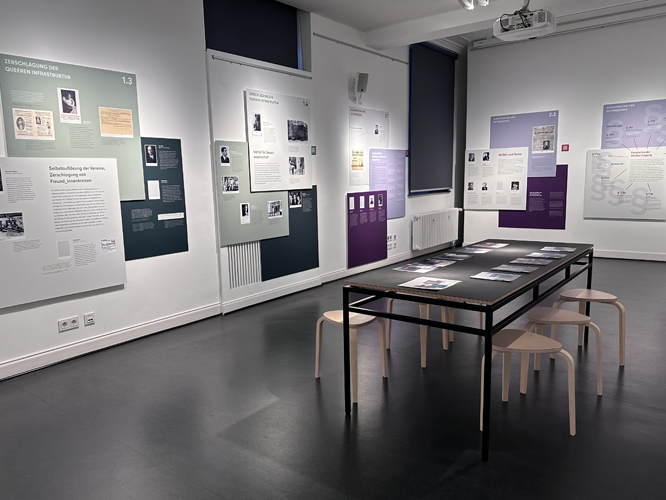 Moderne Ausstellung in einem Museum mit Informationswänden, Fotos, Dokumenten und einem zentralen Tisch mit weiteren Exponaten sowie Sitzgelegenheiten.