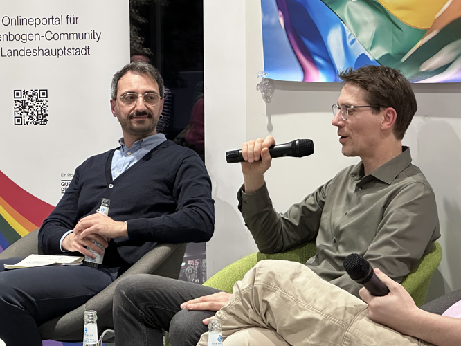 Zwei Männer im Gespräch auf der Bühne mit Mikrofonen, im Hintergrund ein Banner mit der Aufschrift 'Das Onlineportal für die Regenbogen-Community in der Landeshauptstadt'.