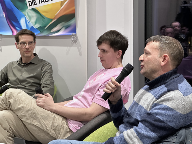 Drei Personen im Gespräch auf der Bühne, eine hält ein Mikrofon, im Hintergrund ein Banner mit Regenbogenfarben und der Aufschrift 'DIE TALKREIHE'.