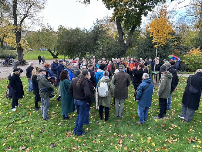 Gruppe von Menschen, die sich im Freien in einem Park versammeln; die Teilnehmer:innen stehen im Kreis auf einer grasbewachsenen Fläche mit herbstlichen Blättern und Bäumen im Hintergrund.