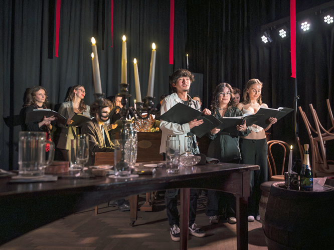 Eine Gruppe von Schauspielenden steht auf einer Bühne und mit Textmappen in der Hand, während im Vordergrund ein Tisch mit Gläsern, Flaschen und Kerzen zu sehen ist.