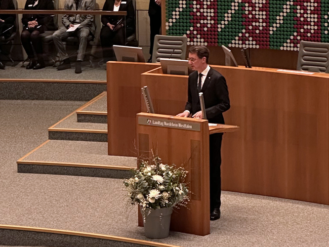 Ein Mann steht an einem Rednerpult mit der Aufschrift "Landtag Nordrhein-Westfalen", vor dem ein Blumenstrauß mit weißen Blüten steht.