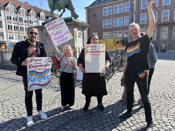 Vier Personen auf einem Platz halten Schilder mit der Aufschrift „Altern unter Regenbogen ist für mich …“ und setzen sich für Respekt und selbstbestimmtes Leben älterer LGBTQ+-Menschen ein.