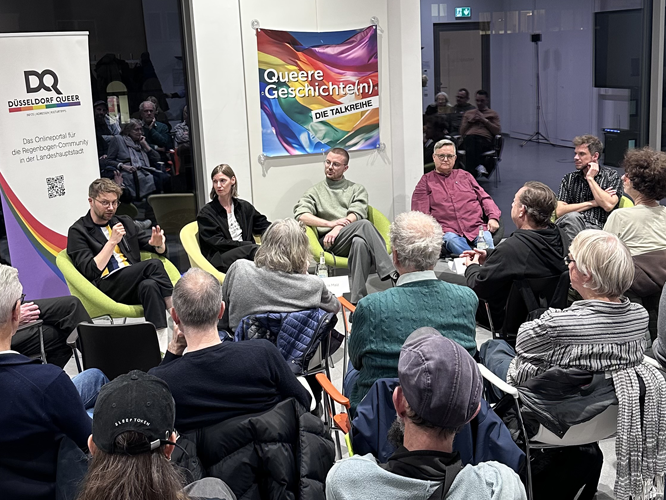 Podiumsdiskussion zu queeren Geschichten: Fünf Personen diskutieren vor Publikum, im Hintergrund ein Banner mit ‚Queere Geschichte(n) – DIE TALKREIHE‘ und ein Logo von ‚Düsseldorf Queer‘.