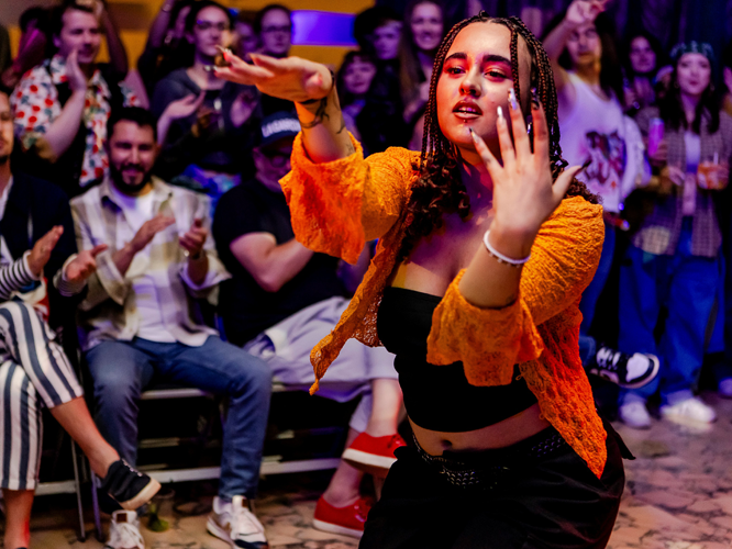 Person in orange-schwarzem Outfit performt selbstbewusst auf der Bühne, während das Publikum im Hintergrund zuschaut.