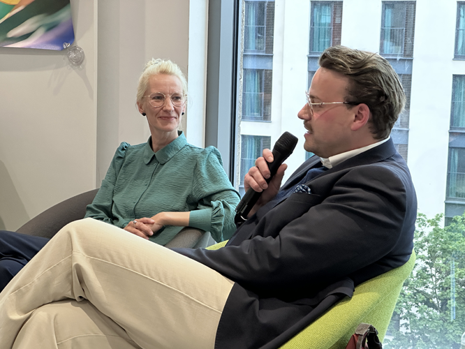 Zwei Personen sitzen auf Sesseln bei einer Podiumsdiskussion: Eine trägt ein grünes Hemd und beige Hose, die andere ein dunkles Jackett und hält ein Mikrofon. Im Hintergrund sind große Fenster mit Blick auf Gebäude und Bäume zu sehen.