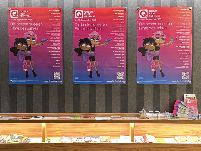 Drei Poster des Queer Film Festivals 2025 mit dem Motto ‚Die besten queeren Filme des Jahres‘ an einer Wand über einem Infotisch mit Flyern und Broschüren