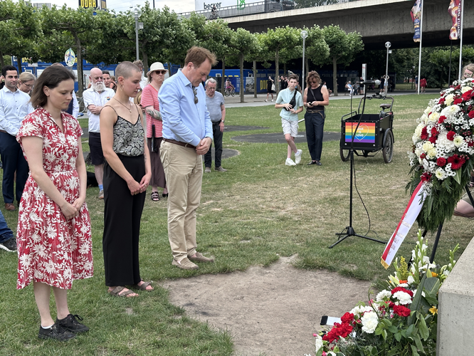 Drei Personen stehen schweigend vor Gedenkort mit Blumenkränzen