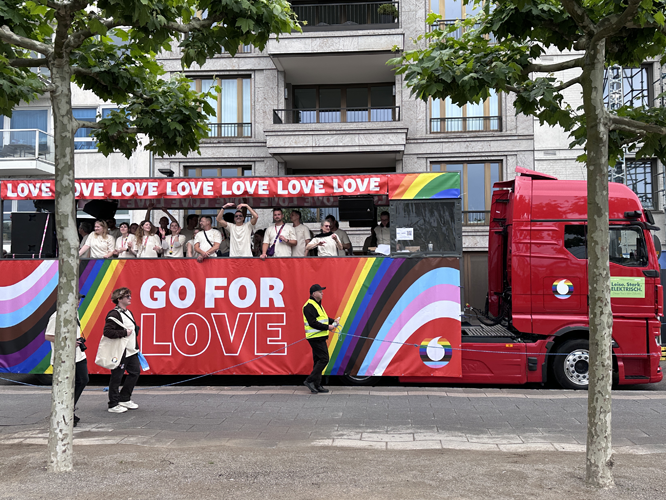 Roter LKW mit Regenbogen-Dekoration und der Aufschrift 'GO FOR LOVE', auf dem Menschen stehen und feiern.