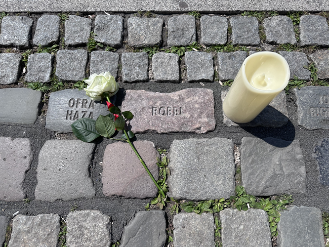 Pflastersteine mit Namen, daneben eine weiße Rose und eine Kerze