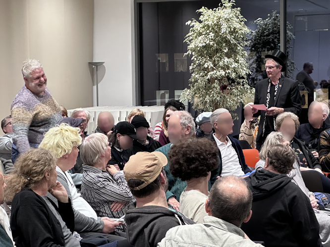 Eine Gruppe von Menschen sitzt in einem Raum und verfolgt aufmerksam eine Darbietung zwischen zwei stehenden Männern. Im Hintergrund steht eine große Pflanze.
