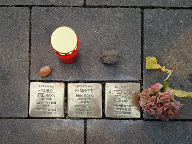 Drei Stolpersteine im Bodenpflaster zur Erinnerung an Emanuel Freimark, Henriette Freimark und Alfred Ludwig Freimark. Die Messingplatten sind mit Namen, Geburts- und Deportationsdaten beschriftet. Daneben liegen eine rote Kerze, Steine und Blumen.