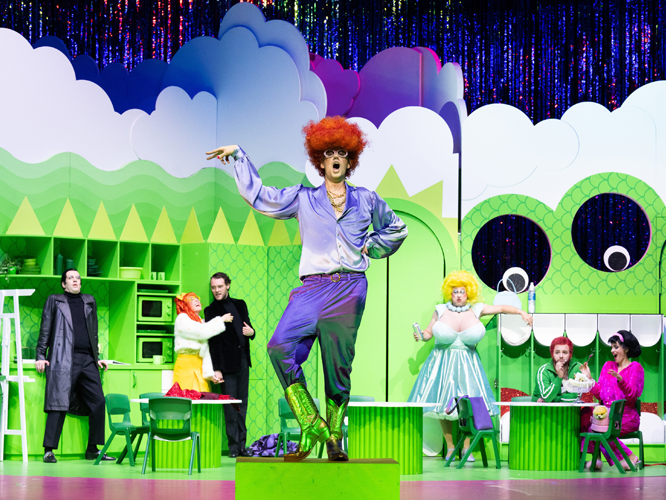 Bunte Theaterszene mit Darstellern in extravaganten Kostümen auf einer grünen Bühne mit übertriebenen Requisiten.