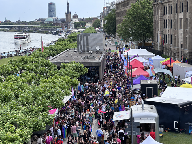 Große Menschenmenge bei einem Pride-Festival an einem Fluss mit bunten Ständen, einer Bühne und historischen Gebäuden im Hintergrund.