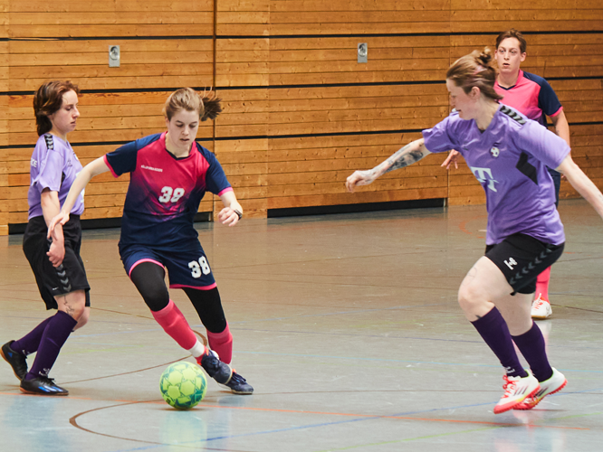 Drei Fußballerinnen in einer Hallensportstätte, die sich aktiv um den Ball duellieren, während eine Spielerin mit der Nummer 38 den Ball kontrolliert.