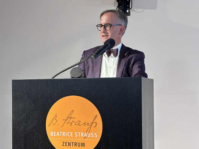 Ein Mann (Helmut Metzner) in einem violetten Anzug und Brille hält eine Rede an einem Pult mit dem Logo und der Aufschrift 'Beatrice Strauß Zentrum'.