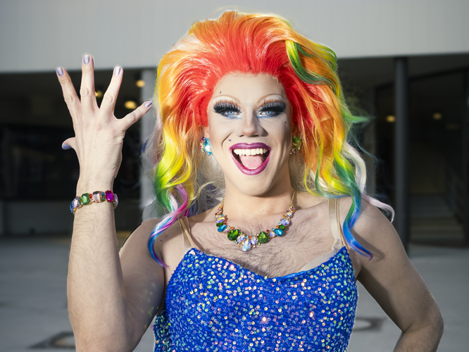 Eine Person mit buntem, rainbow-farbenem Haar und auffälligem Make-up trägt ein blaues, glitzerndes Kleid und posiert mit einer Handgeste vor einem neutralen Hintergrund.