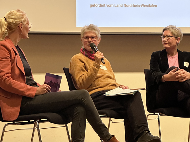 Drei Frauen sitzen auf einer Bühne bei einer Podiumsdiskussion; die mittlere Frau spricht in ein Mikrofon, während die anderen beiden zuhören. Im Hintergrund ist eine Leinwand mit dem Text 'gefordert vom Land Nordrhein-Westfalen' zu sehen.
