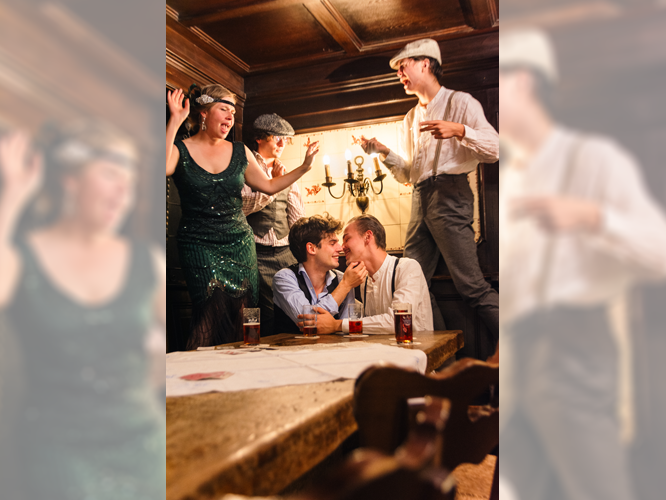 In einer urigen Gaststube sitzen zwei junge Männer, die sich küssen. Um sie herum tanzen Personen auf debn Bänken. Auf dem Tisch stehen Gläser mit Altbier.
