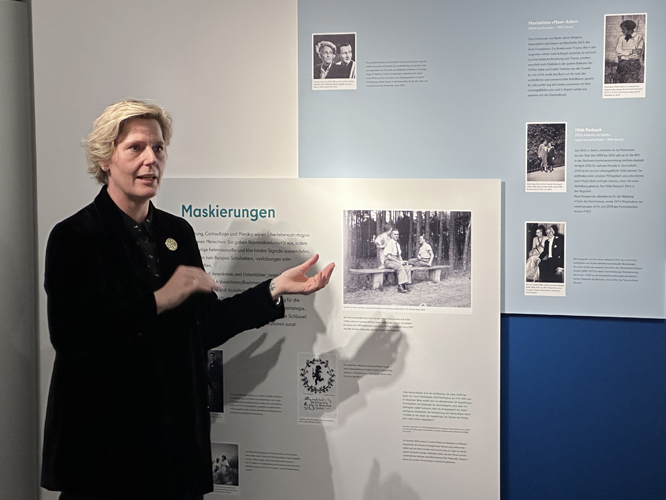 Eine Person (Astrid Hirsch-von Borries) hält einen Vortrag vor einer Ausstellungstafel mit historischen Fotos und Informationen zum Thema 'Maskierungen'.
