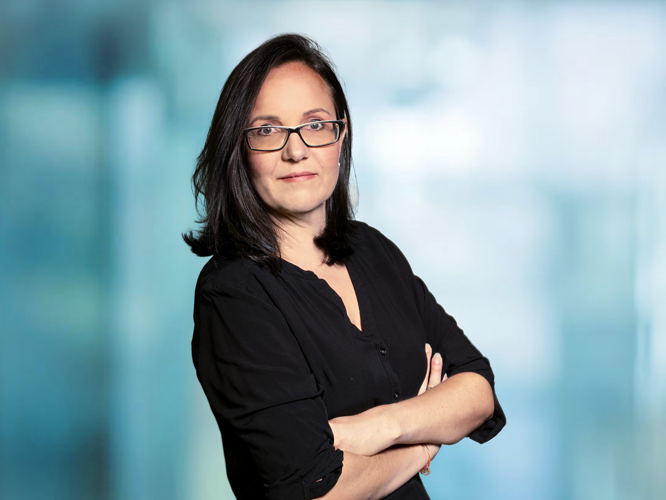 Porträt einer Person mit kurzen dunklen Haaren und Brille, die selbstbewusst in die Kamera blickt und die Arme verschränkt hat, vor neutralem Hintergrund.