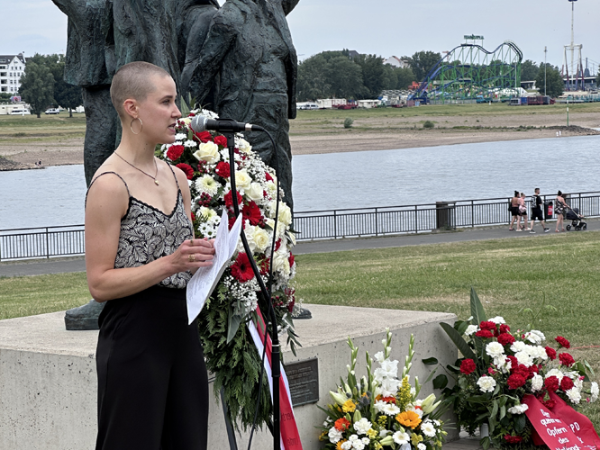 Frau hält Rede mit Blumenkranz vor Denkmal am Fluss