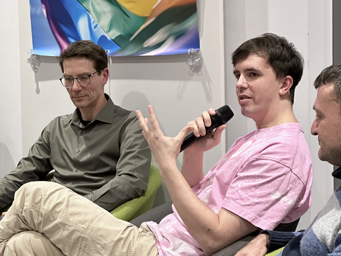 Drei Personen im Gespräch auf bequemen Sesseln, eine Person hält ein Mikrofon und gestikuliert während der Diskussion, im Hintergrund ein Regenbogen-Banner.