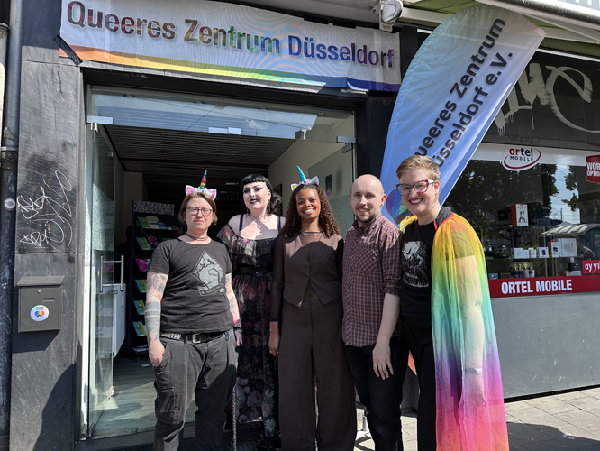 Fünf Personen stehen vor dem Eingang des Queeres Zentrum Düsseldorf, zwei tragen Katzenohren, eine eine Regenbogenflagge als Cape. Über dem Eingang hängt ein Banner mit dem Namen des Zentrums.