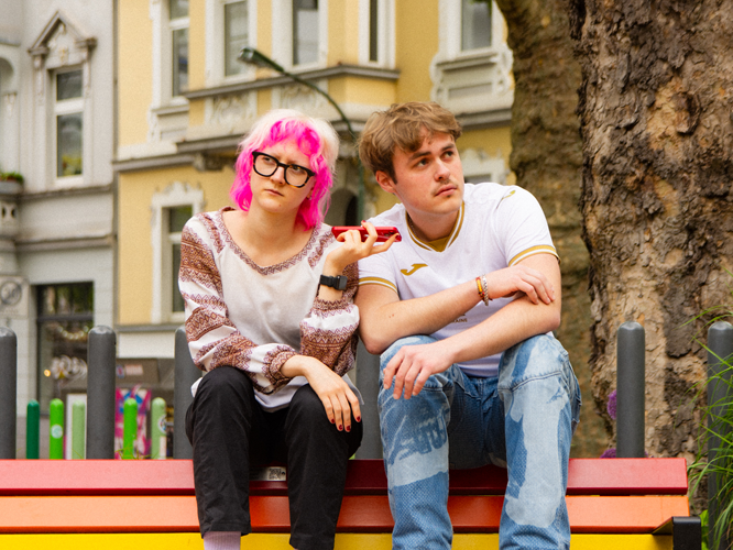 Zwei Personen sitzen auf einer bunten Bank: Eine Frau mit pinken Haaren, Brille und gestreiftem Pullover hält ein Smartphone, während ein Mann in weißem T-Shirt und Jeans entspannt daneben sitzt. Im Hintergrund sind gelbe Gebäude und ein Baum zu sehen.