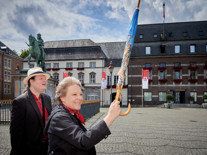 Zwei lächelnde Personen stehen auf einem gepflasterten Platz vor einem historischen Gebäude mit Flaggen; die Person im Vordergrund hält einen bunten Regenschirm in die Höhe, während im Hintergrund eine Reiterstatue zu sehen ist.