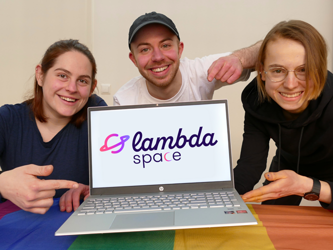 Drei lächelnde Personen sitzen gemeinsam an einem Tisch und präsentieren stolz einen Laptop mit dem Logo von Lambda Space, während eine Regenbogenflagge im Vordergrund liegt.