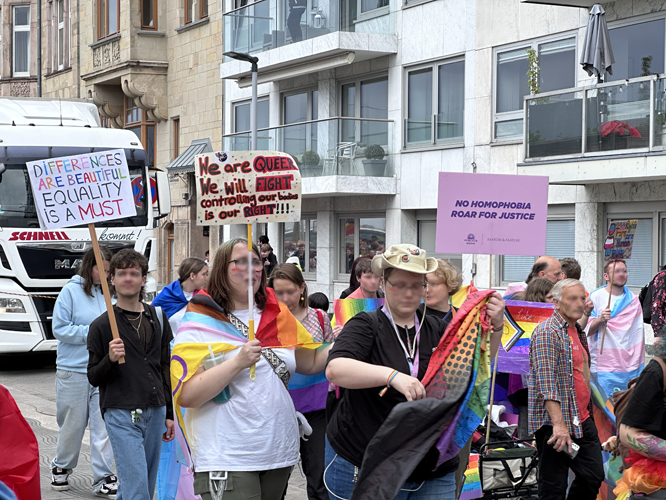Pride-Demonstration mit Teilnehmenden, die Schilder mit Botschaften wie 'Differences are beautiful, equality is a must' und 'No homophobia – roar for justice' halten.