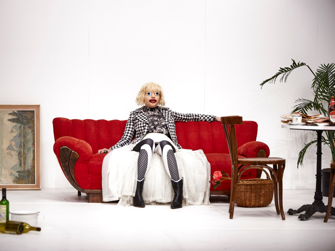 Person in Clowns-Make-up mit kariertem Oberteil und weißem Umhang sitzt auf einem roten Sofa in einem ungewöhnlichen, künstlerisch gestalteten Raum.