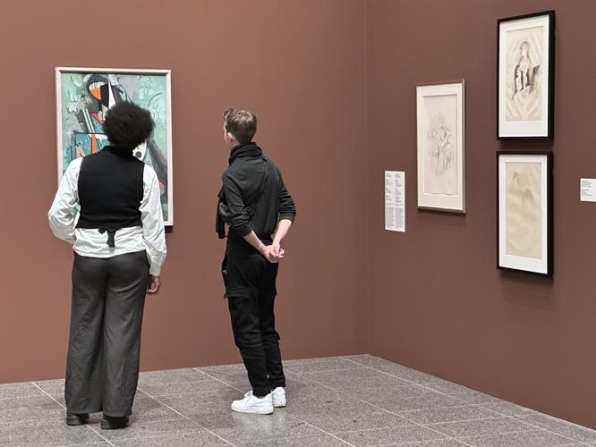 Zwei Personen betrachten in einer Ausstellung ein abstraktes Gemälde an einer braunen Wand, daneben hängen drei Zeichnungen in Rahmen.