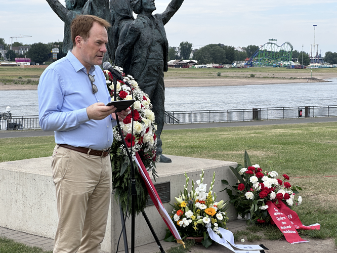 Mann hält Rede vor Denkmal mit Blumenkränzen am Rheinufer