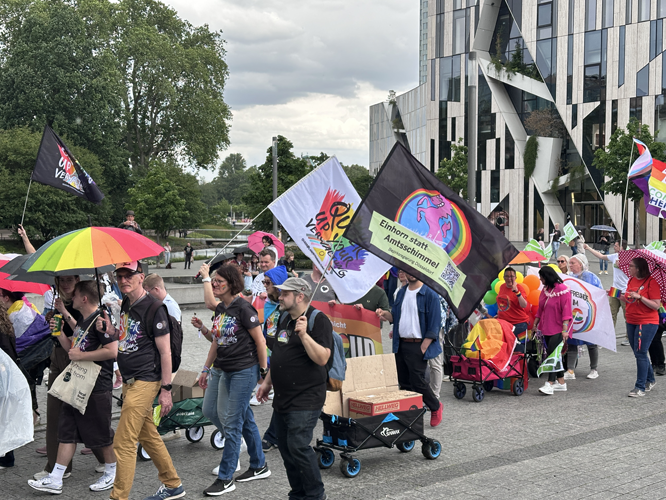 Pride-Parade mit bunt gekleideten Menschen, Regenbogenfahnen und einem Banner mit der Aufschrift 'Einhorn statt Amtschimmel'.