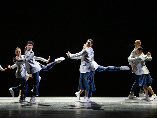 Balletttänzer*innen mit blauen Hosen, grauen Jackets und weißen Turnschuhen. Miteinander tanzend und zum Teil mit hochgestreckten Beinen.