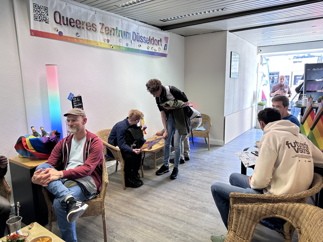 nnenraum des Queeres Zentrum Düsseldorf mit mehreren Personen, die in gemütlicher Atmosphäre auf Flechtstühlen sitzen oder stehen. An der Wand hängt ein Banner mit dem Namen des Zentrums.