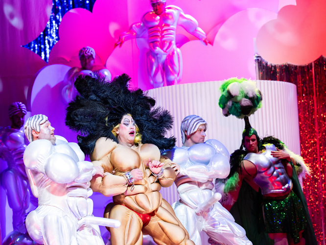 Bunte Bühnenszene mit Performern in extravaganten Kostümen, darunter ein Künstler mit übertriebenen Muskelattrappen und einem großen Federkostüm, umgeben von weiteren Darstellern in fantasievollen Outfits und glitzernder Bühnenausstattung.