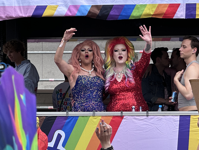Zwei Personen in glitzernden Outfits und bunten Perücken auf einem Wagen mit Regenbogen-Dekoration, die jubelnd ihre Arme heben.