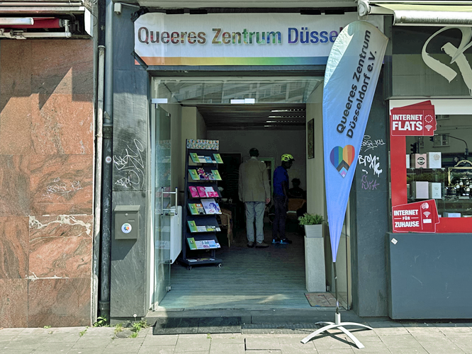 Eingang des Queeren Zentrums Düsseldorf mit blauer Flagge und Regalständer mit Flyern. Im Hintergrund sind Personen zu sehen. Rechts ein benachbartes Schaufenster mit Internet-Werbung, links eine Hauswand mit Graffiti.