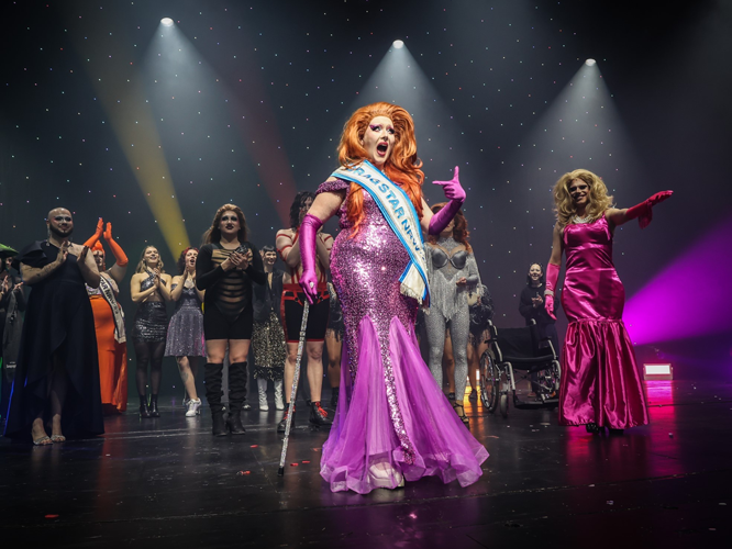 Gewinner:in einer Drag-Show in glitzerndem lila Abendkleid und mit Schärpe auf der Bühne, umgeben von jubelndem Publikum.