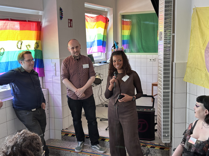 Drei Personen stehen in einem Raum des Queeres Zentrum Düsseldorf, eine hält ein Mikrofon. Im Hintergrund hängen Regenbogenflaggen und bunte Banner.