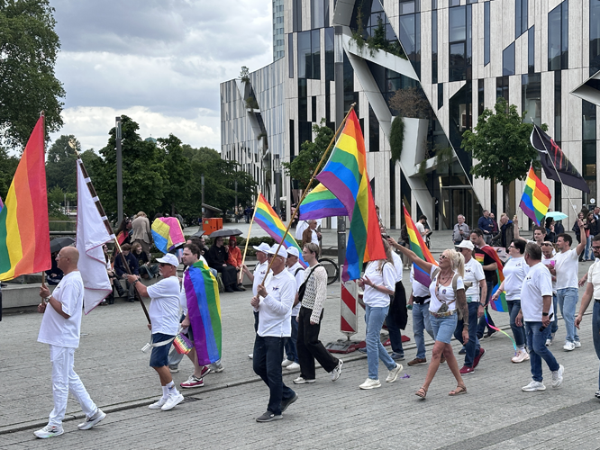 Pride-Demonstration mit Menschen in Weiß, die Regenbogenfahnen tragen und durch eine Stadt marschieren.