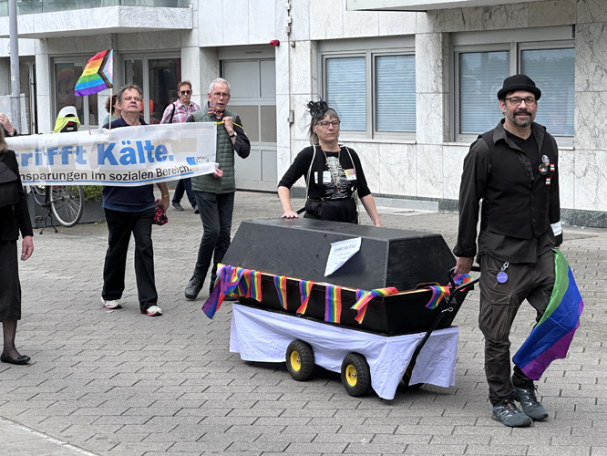 Gruppe von Menschen bei einer Pride-Demonstration, die einen Sarg mit Regenbogen-Schleifen ziehen und ein Banner mit der Aufschrift 'Nähe trifft Kälte. Gegen Einsparungen im sozialen Bereich' tragen.