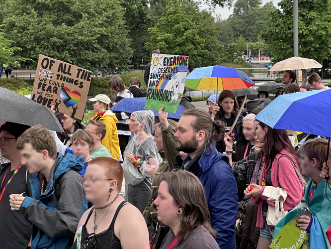 Regenbogen-Parade mit Menschen, die bunte Schilder und Regenschirme halten, darunter eines mit der Aufschrift 'Of all the things we could fear, we pick love'.