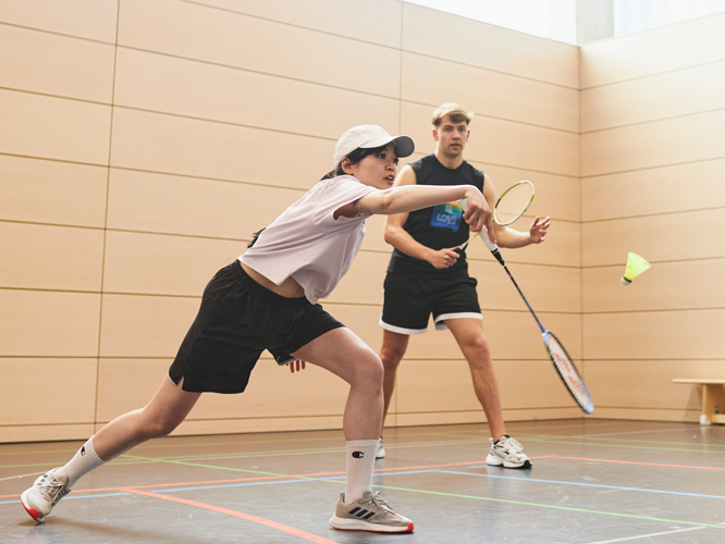 Zwei Personen spielen Badminton in einer Sporthalle, eine Person im Vordergrund schlägt den Federball mit dem Schläger.